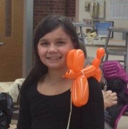 A girl smiles while holding a bright orange balloon animal shaped like a dog