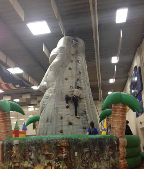 A child climbs a large inflatable rock wall surrounded by palm trees in an indoor setting