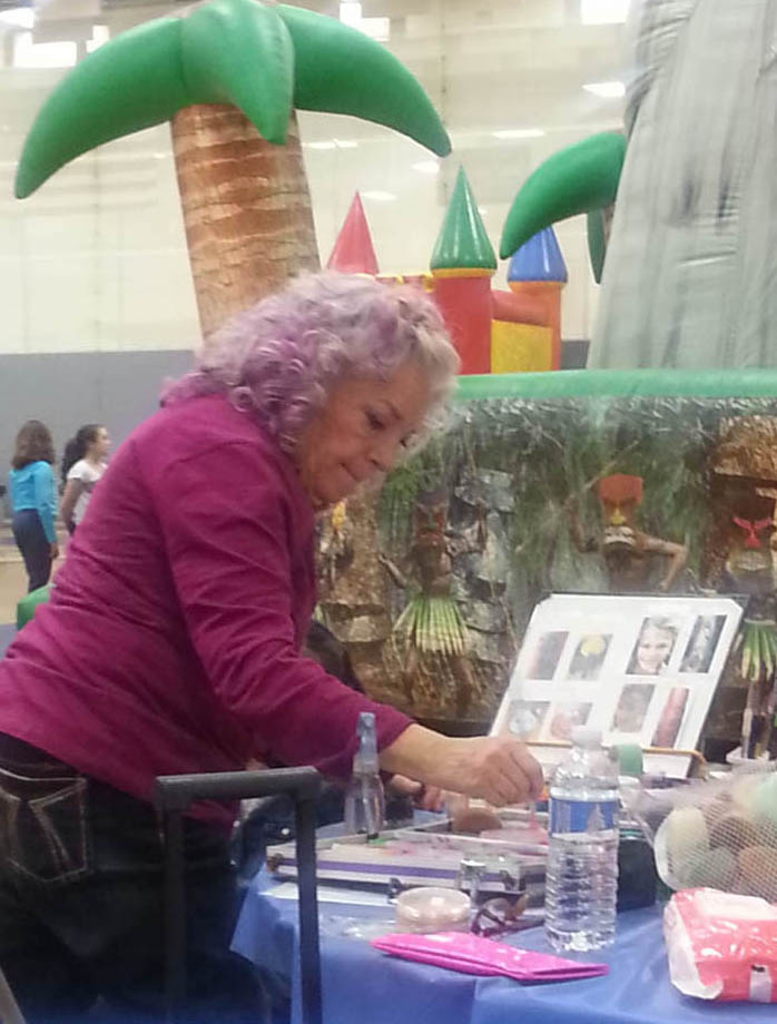 A woman with curly purple hair applies paint at a decorated table